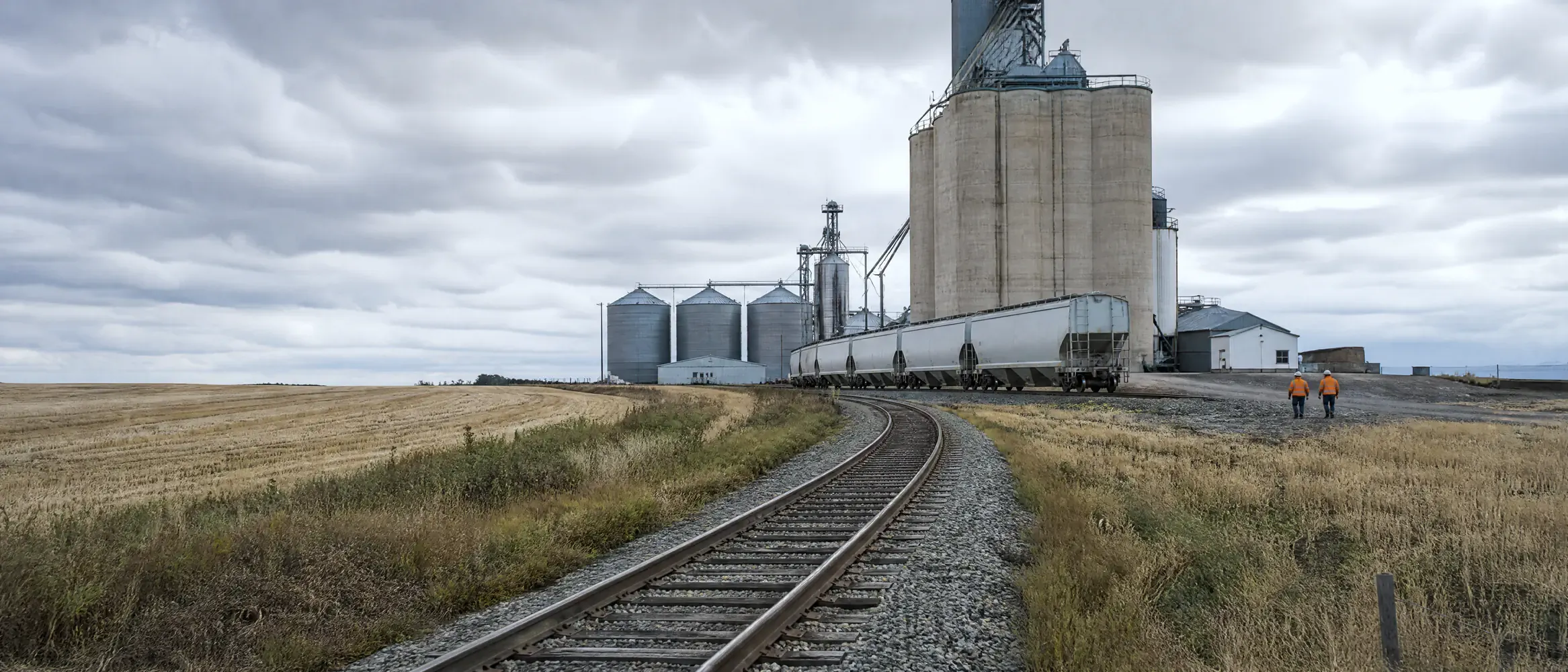 Industrial spur curving across Alberta prairie fields to a grain elevator with covered hoppers