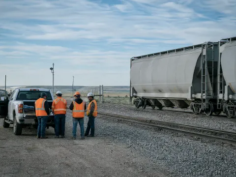 Four-person crew holding a tailgate safety meeting beside covered hoppers on a prairie siding
